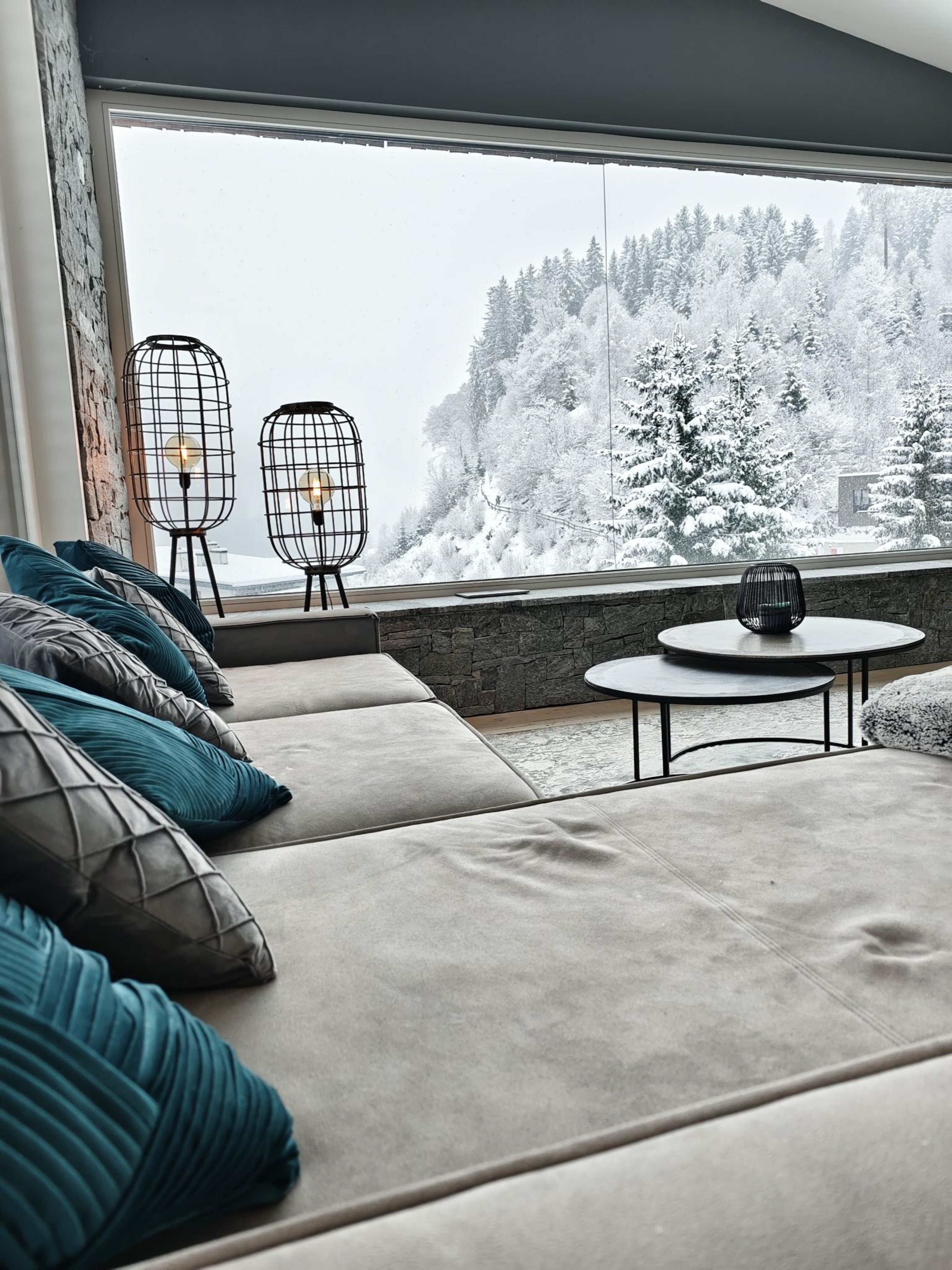 Großes hellgraues Sofa mit blauen und grauen Kissen vor Panoramafenster mit Blick auf schneebedeckte Tannen im Luxus Chalet in Wagrain.
