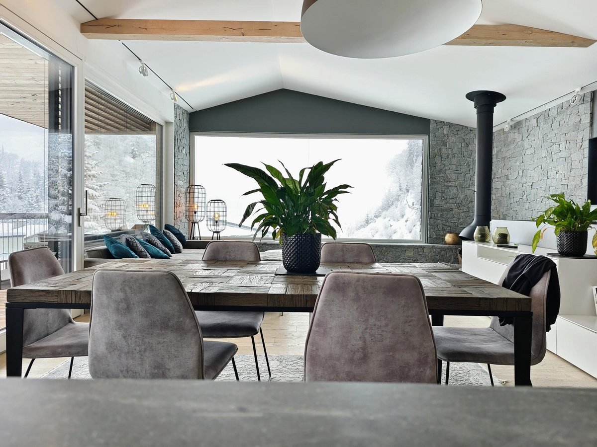 Esszimmer mit rustikalem Holztisch, sechs grauen Stühlen, großen Fenstern mit Blick auf verschneite Berge und Kaminofen in einem Luxus-Chalet in Wagrain.