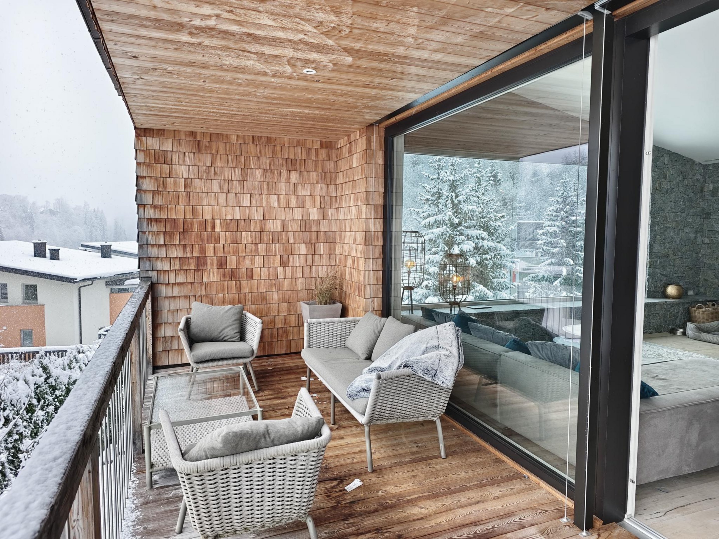 Balkon mit Holzboden und Holzdecke, drei gepolsterte Stühle und ein Sofa, dahinter große Glasschiebetür zu einem Schlafzimmer. Luxus Chalet Wagrain