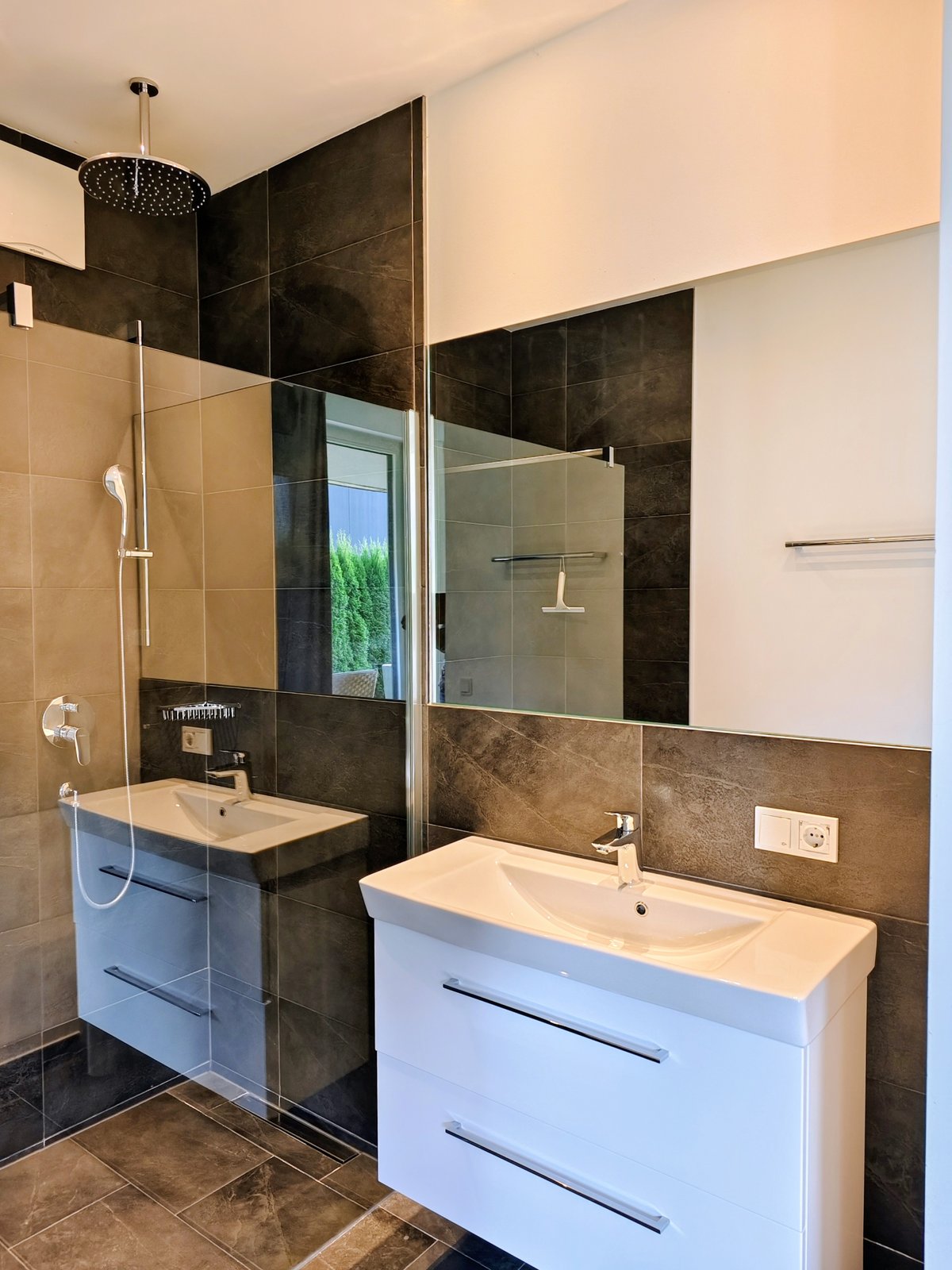 Modernes Badezimmer im Chalet in Wagrain mit weißem Waschbecken, großem Spiegel und begehbarer Dusche mit Regenduschkopf.